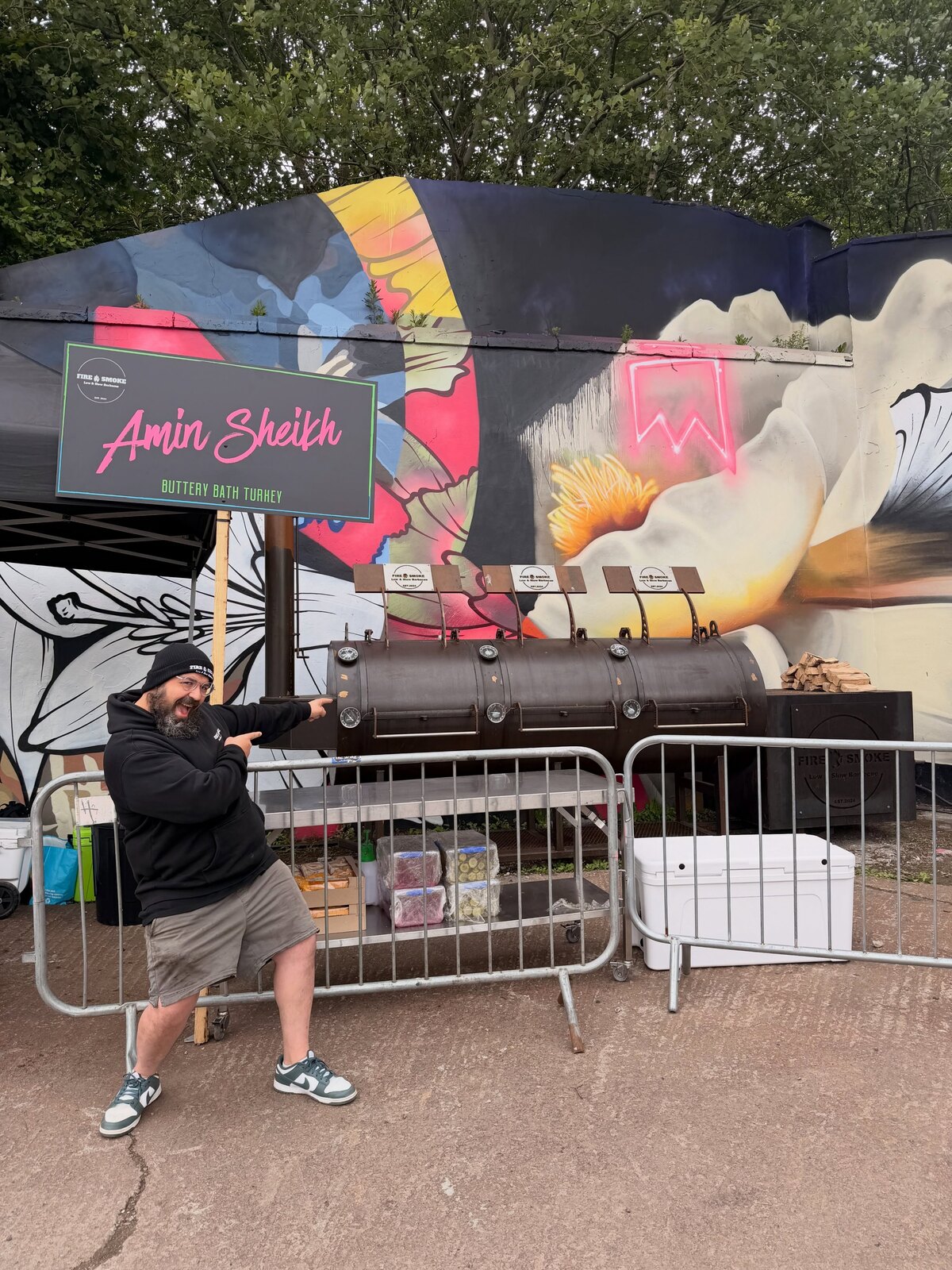 Pitmaster Amin Sheikh pointing at his offset smoker beside a Fire & Smoke sign at a street-food event